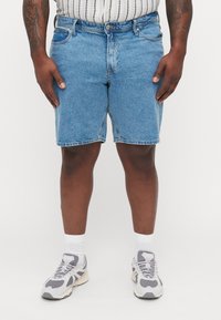 Pantalones cortos de mezclilla en un lavado azul claro con un corte holgado, que cuentan con cinco bolsillos, una cremallera y costuras visibles. Llevados con zapatillas grises y calcetines blancos.