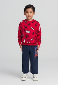 Felpa rossa con grafiche nere Marvel, abbinata a pantaloni blu con design di Spider-Man. Bambino che indossa scarpe bianche.