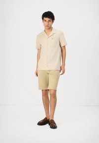 Lys beige kortermet skjorte med krage og frontlomme, kombinert med khaki-shorts. Mørkebrune loafers fullfører antrekket.