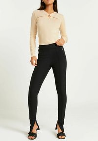 Beige Langarmoberteil mit einem runden Ausschnitt an der Brust; kombiniert mit schwarzen, eng anliegenden Hosen mit Reißverschlusstaschen und seitlichen Schlitzen am Saum.
