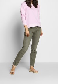 Femme portant un haut à manches longues rose clair, un pantalon slim olive, et des sandales à bout ouvert beiges sur un fond blanc uni.