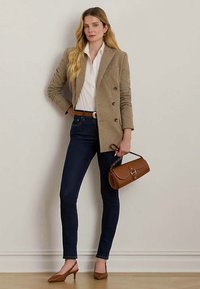 Ženska v bež karo blazerju, beli bluzici, temnih kavbojkah, rjavem pasu, rjavih petah, drži rjav nahrbtnik, stoji ob navadni steni.