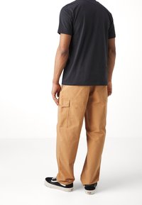 Ciemnoszara koszulka z krótkim rękawem w połączeniu z beżowymi spodniami cargo z bocznymi kieszeniami. Model stoi na białym tle, nosząc czarne trampki.