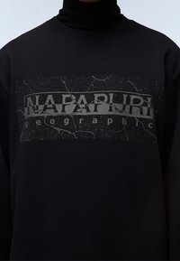 Sudadera negra con un logotipo rectangular texturizado agrietado que muestra "NAPAPIJRI geographic" en el pecho.