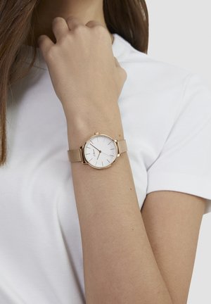 Rond horloge met een witte wijzerplaat en gouden accenten, voorzien van een mesh band. Het horloge wordt gedragen om een pols tegen een effen witte achtergrond.