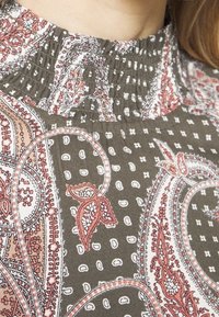 Tissu gris avec un motif paisley agrémenté de accents rouges et blancs. L'encolure présente des détails smockés et de petits pois blancs sur l'ensemble.