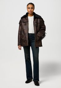 Brown leather jacket with fringe accents, black faux fur collar, zip front, paired with a white henley shirt and dark blue fitted jeans.