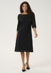 Black lace dress with three-quarter sleeves, floral pattern, scalloped hem, and a relaxed fit, paired with black high-heeled shoes.