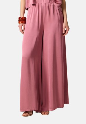 Femme portant un pantalon rose ample à jambes larges, associé à des sandales ouvertes marron et deux bracelets marron épais au poignet gauche.