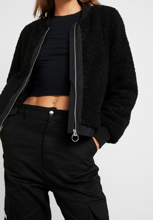 Vrouw die een zwart cropped rib top draagt, een gestructureerd zwarte jas met rits en zwarte broek met cargozakken, handen in de zakken.