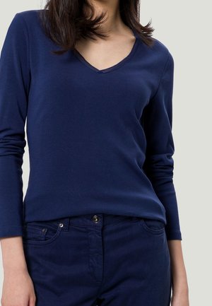 Frau trägt ein dunkelblaues Langarmshirt mit V-Ausschnitt und passende dunkelblaue Hose vor hellem Hintergrund.