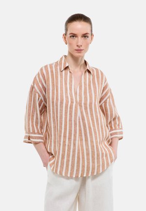 Femme portant une chemise ample à rayures verticales beige et blanc avec les manches retroussées et un pantalon blanc, debout les mains dans les poches.