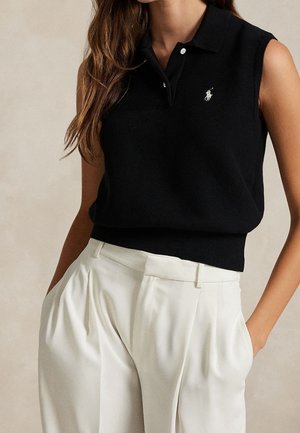 Nainen pukeutunut hiatttomaan mustaan neulottuun poolopaitaan, jossa on logo, ja valkoisiin korkeavyötäröisiin laskostettuihin housuihin, kädet taskuissa, neutraalia taustaa vasten.