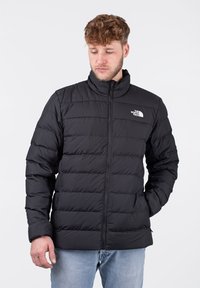 Giacca nera imbottita con colletto alto, chiusura con zip e logo bianco. Presenta un design trapuntato e maniche lunghe con polsini a maglia.