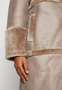 Long faux fur coat in soft taupe, featuring plush cuffs, a textured body, and a side pocket. Smooth and warm material.