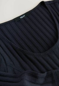 Donker ribstof met een ronde halslijn, met een label dat "NEXT" zegt. Het kledingstuk is voorzichtig gevouwen tegen een neutrale achtergrond.