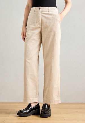 Partie inférieure portant un pantalon large en velours côtelé beige et des mocassins compensés noirs, se tenant sur un sol en bois devant un mur neutre.