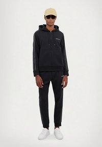 Svart hettejakke med glidelås og snorer, sidestriper, kombinert med matchende svarte joggebukser og hvite joggesko. Tilbehør inkluderer en beige caps og solbriller.