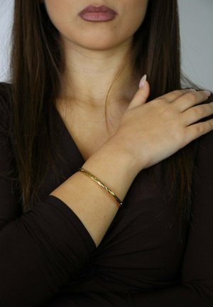 Vrouw in donkerbruine top, die haar linkerschouder vasthoudt met haar rechterhand, gouden gedraaide armband zichtbaar aan haar linkerpols.