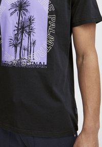 Camiseta de algodón negra con un gráfico púrpura de palmeras, y el texto "BLEND ORIGINAL" en blanco. Corte regular, textura suave.