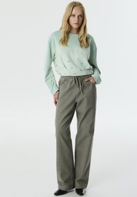 Sudadera verde claro con bordado sutil, combinada con pantalones de pierna ancha grises y texturizados; ambos ofrecen un ajuste relajado y casual. Unos zapatos negros completan el conjunto.