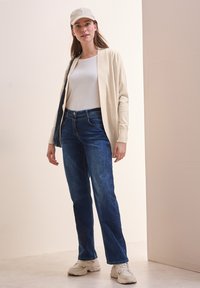 Beige cardigan over een wit shirt, gecombineerd met blauwe jeans en witte sneakers. De cardigan heeft geribbelde randen en een ontspannen pasvorm.