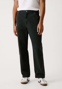 Schwarze Baumwollhosen mit geradem Schnitt, ausgestattet mit Vorderaschen und einem Knopfverschluss. Kombiniert mit weißen Sneakers, Aufnahme von der Taille abwärts.
