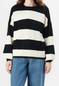 Schwarz-cream gestreifter Pullover mit gerippter Textur, Rundhalsausschnitt und übergroßen Ärmeln, kombiniert mit blauen Jeans.