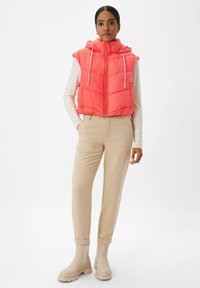 Padded sleeveless vest w jaskrawym koralowym kolorze z przednim zamkiem, regulowanym kapturem i białymi sznurkami, w połączeniu z beżowymi spodniami i botkami do kostek.