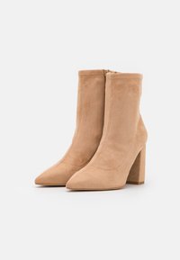 Bottines beiges en daim avec un bout pointu et un talon épais. Elles comportent une fermeture éclair latérale et une texture lisse, avec des détails de couture minimalistes.