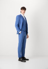 Modra trodelna obleka z fitted jakno in ravnimi hlačami, kombinirana z belo srajco in črnimi vezalkami.