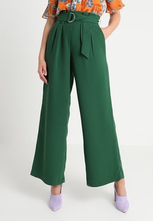 Vrouw draagt hoge taille wijde groene broek met een stoffen riem en zilveren gesp, gecombineerd met paarse hakken.