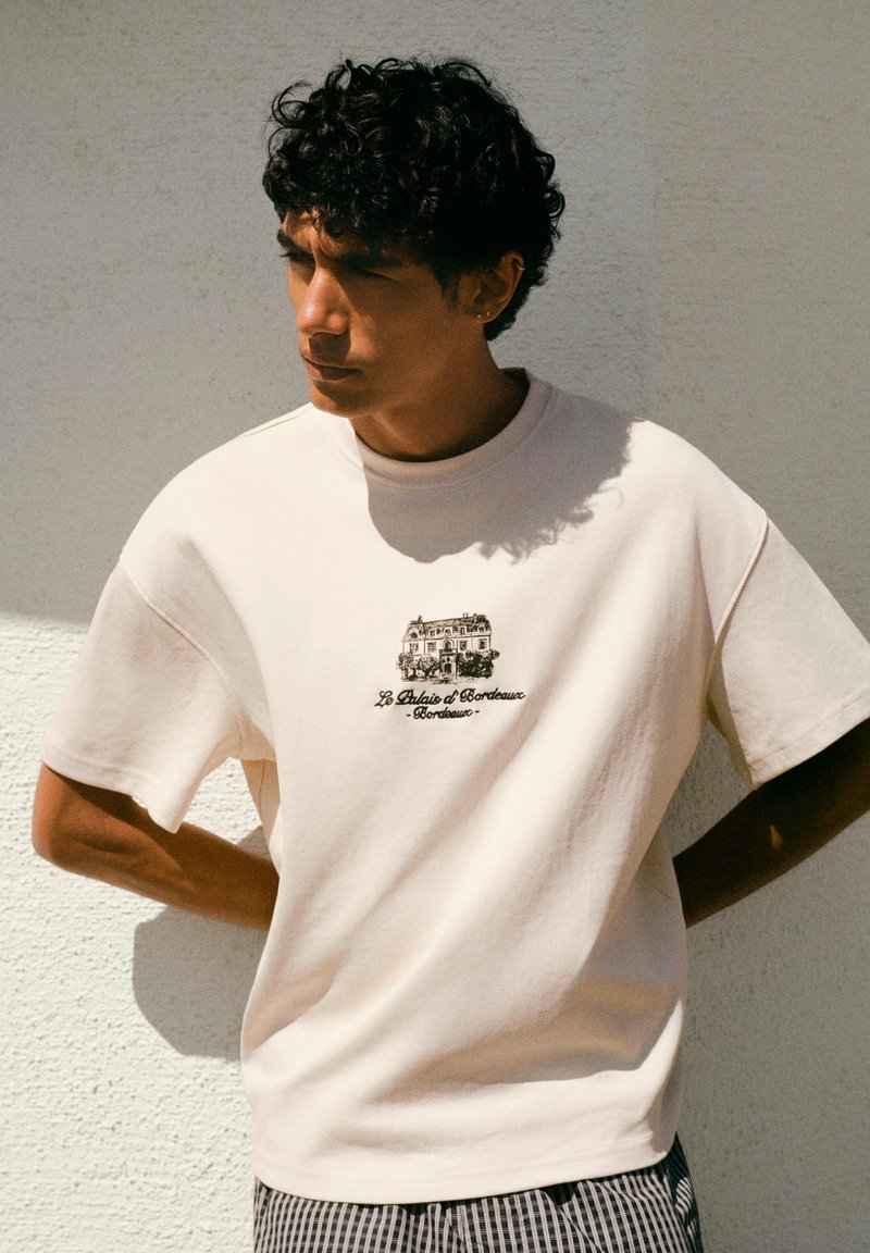 Jeune homme aux cheveux bouclés portant un t-shirt écru avec un petit croquis noir et le texte "Le Palais d'Bordeaux" sur la poitrine, debout contre un mur.