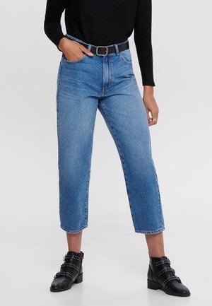 Kobieta ubrana w niebieskie dżinsy z wysokim stanem i skróconą długością, czarny pasek, czarną bluzkę z długim rękawem oraz czarne botki z ćwiekami, stojąca z jedną ręką w kieszeni.