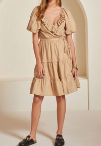 Beige linnen wikkeljurk met pofmouwen, ruches langs de halslijn en een gelaagde rok. Gestyleerd met zwarte instapschoenen.