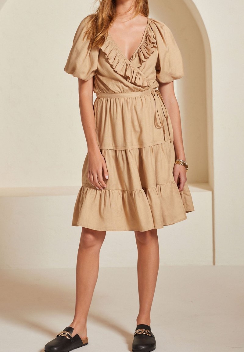 Beige linnen wikkeljurk met pofmouwen, ruches langs de halslijn en een gelaagde rok. Gestyleerd met zwarte instapschoenen.