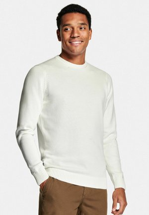 Lächelnder Mann, der einen cremefarbenen Strickpullover und braune Hosen trägt, mit einer Hand in der Tasche stehend und seitlich auf weißem Hintergrund blickend.