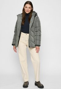 Woman wearing a grey puffer jacket over a black turtleneck, cream pants, and dark green chunky boots standing against a plain background.