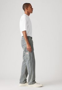Camisa blanca de manga corta y pantalones de pierna ancha a rayas grises con parches. Calzado: zapatillas blancas. Vista de perfil lateral contra un fondo simple.