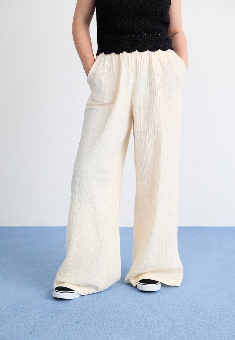 Cream-colored wide-leg pants with textured fabric, featuring front pockets and a relaxed fit, paired with black and white sneakers.