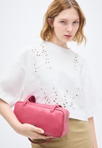 Νεαρή γυναίκα με κοντά ξανθά μαλλιά που φοράει λευκό τοπ με κοψίματα και μπεζ φούστα, κρατώντας μια ορθογώνια ροζ δερμάτινη τσάντα με χερούλια.