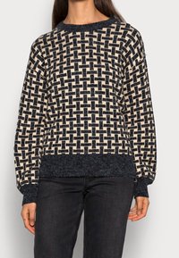 Pull en tricot avec un motif entrelacé crème et bleu foncé, col rond, ourlet texturé et manches longues. Fabriqué en tissu doux.