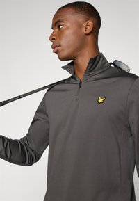 Mørkegrå kvart-zip trøje lavet af glat stof, med et lille gult broderet logo på brystet og en golfklub hvilende på skulderen.
