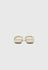 Beige flache Sandalen mit zwei oberen Riemen, einer glatten Textur und einer schwarzen Sohle. Minimalistisches Design mit klaren Linien und unauffälligen Details.