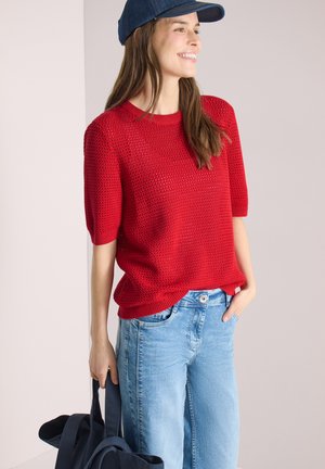 Smiling woman wearing a blue cap, red knit short-sleeve sweater, light blue jeans, holding a navy bag in one hand, hand in pocket.