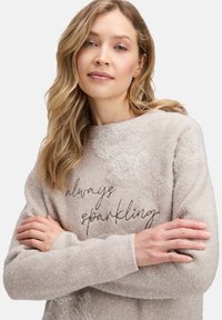 Jasno beżowy sweter wykonany z miękkiego, teksturowanego materiału. Posiada okrągły dekolt oraz napis "zawsze błyszczący" w metalicznej haftowanej aplikacji.