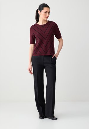 Vrouw met donker haar in een paardenstaart, gekleed in een bordeauxrood topje met korte mouwen en zwarte V-strepen, en zwarte rechte broek, staand met één hand in haar zak.