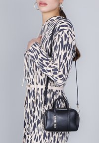 Borsa a mano in pelle nera con chiusura zip e doppio manico, accentuata da dettagli in metallo argentato. Indossata insieme a un vestito a fantasia crema e blu.