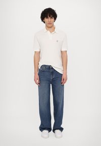 Hvid kortærmet polo med et lille broderet logo, kombineret med løse blå jeans og hvide sneakers. Simpelt design, afslappet pasform.