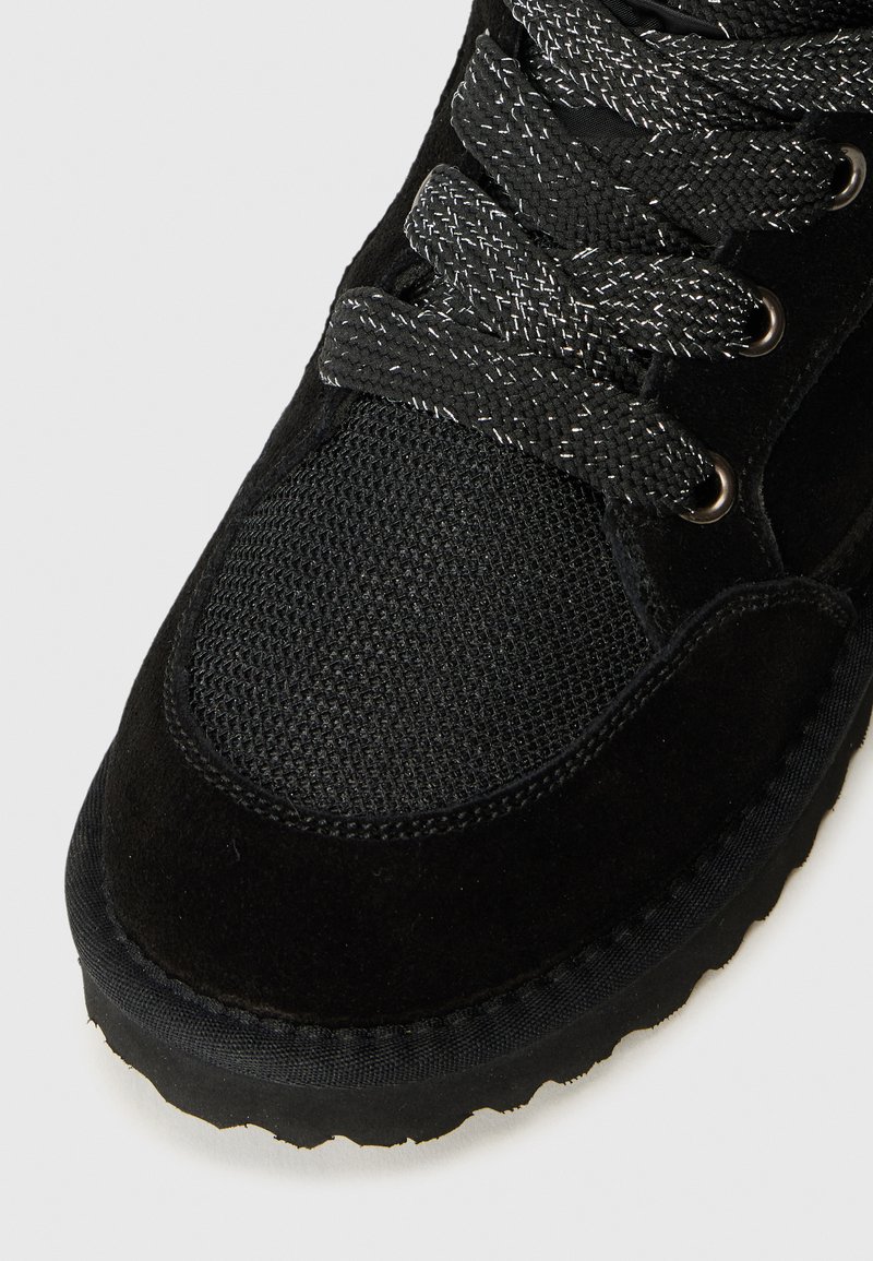 Sneaker en suède noir avec un panneau en maille texturée, des lacets noirs brillants et une semelle en caoutchouc avec un motif de bande de roulement nervuré.